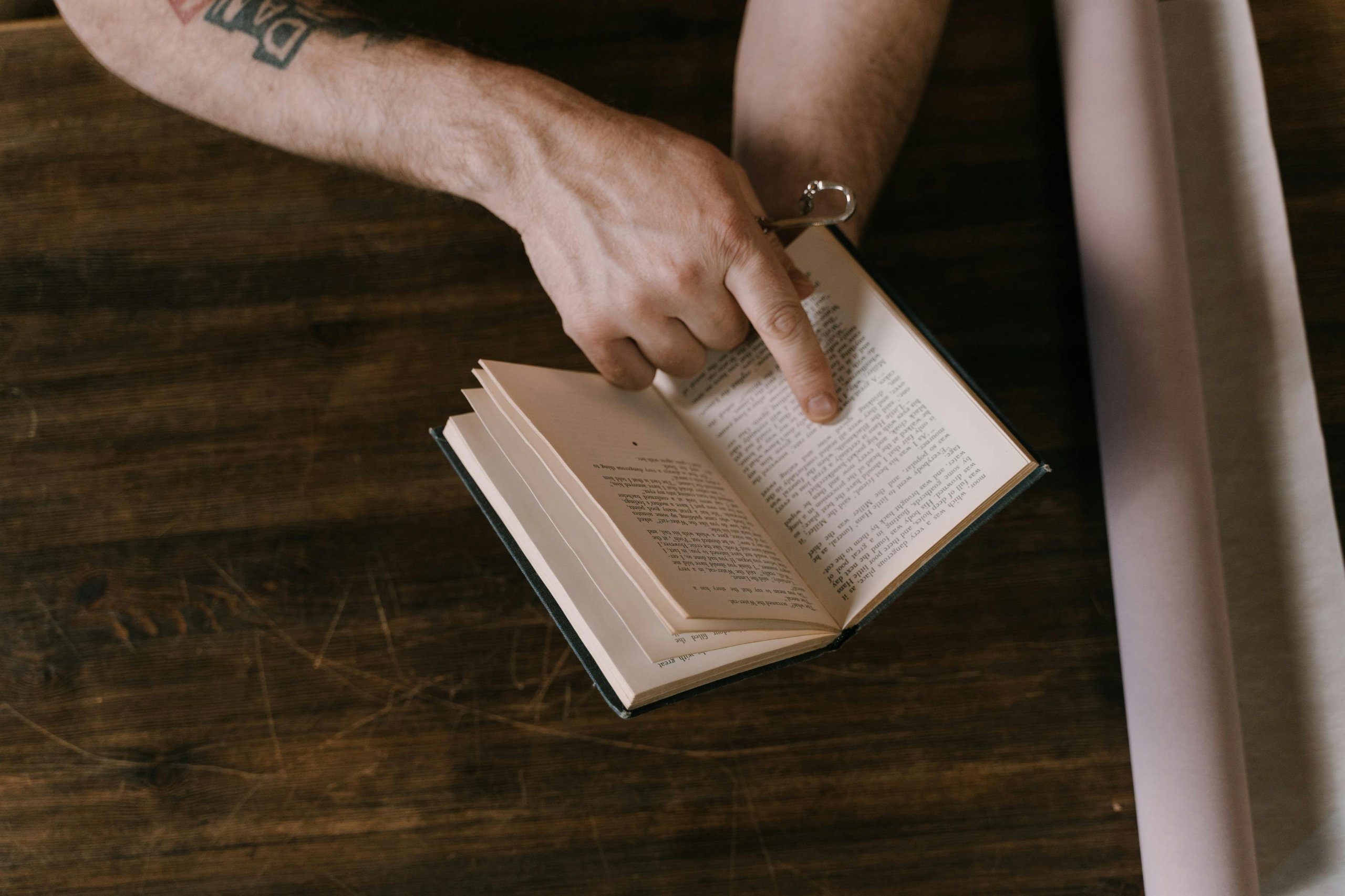 A close-up photographic-style image of an open book resting on a wooden surface (table, desk, or floor — anything with grain). The page is visible but the text is intentionally indistinct (low resolution, slight blur, or pencil-shadow). A single finger rests mid-page — as if holding a line, considering it, or about to underline it. The book is clearly well-handled: worn corners, soft spine, possibly a marginal pencil mark visible. Soft natural side-light from a window. No face. No clock or other context-clichés. The mood captures *re-reading*, the live-thinking quality of returning to a passage that has not yet finished working.